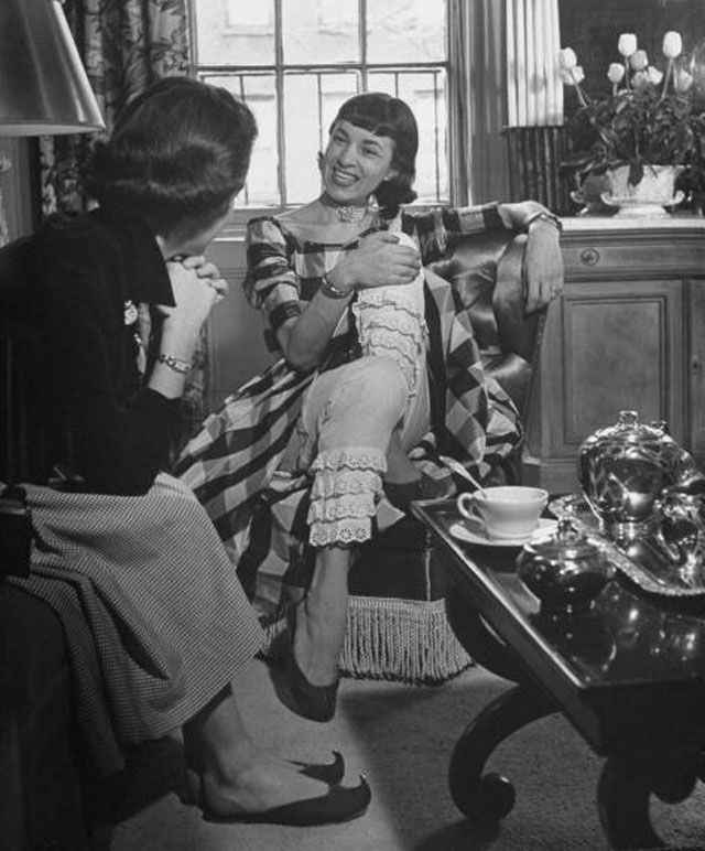 "Designer Anne Fogarty, dressed in plaid taffeta outfit with white lace pantaloons while enjoying a visit from a friend in her apartment."